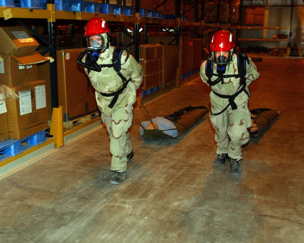 Zwei Militärangehörige in Gasmasken und roten Helmen tragen eine Trage durch ein Lager voller Kartonagen und gelagerter Gegenstände.