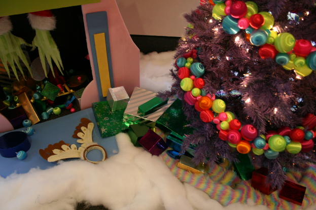 Drinnen steht ein geschmückter Weihnachtsbaum mit Lichterkette und Schmuck rechts, mit Geschenkboxen und Dekorationen in der Mitte, eine Hütte mit Dekorationen oben links und Watte und Wolle unten.