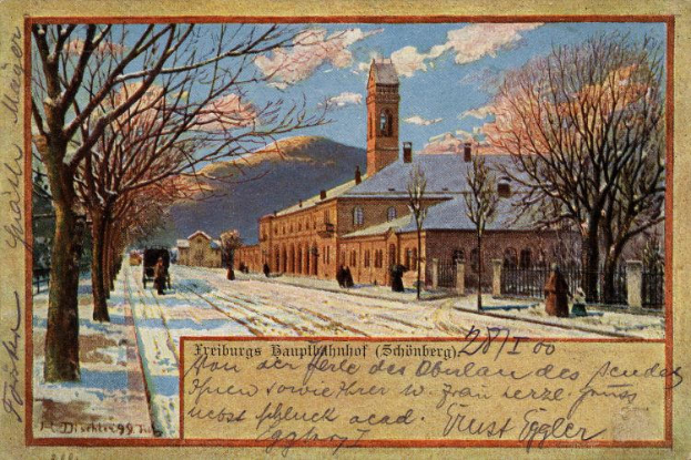 Eine alte Ansichtskarte zeigt ein schneebedecktes Gebäude umgeben von Bäumen, Menschen und einem Zaun, mit einem bewölkten Himmel und der Aufschrift "Freiburg Hauptbahnhof" unten.