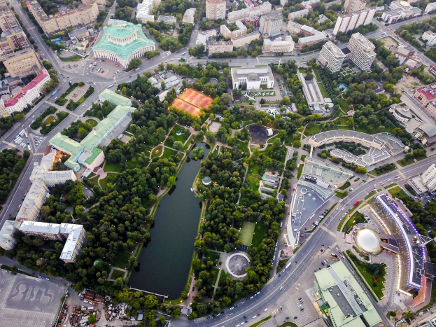 Eine Luftaufnahme einer Stadt mit einem zentralen Park, umgeben von Grünflächen, Gebäuden, Straßen, Fahrzeugen und Wasser.