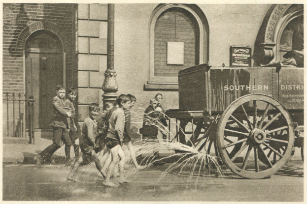 Schwarzes und wei├čes Foto von Kindern, die in der N├Ąhe eines Feuerhydranten vor einem Geb├Ąude mit Fenstern, einer Tür, einem Pfosten und einem Zaun spielen, mit einem Wagen im Hintergrund.