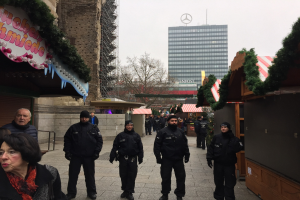 Polizeibeamte in schwarzen Uniformen stehen vor einem geschmückten Weihnachtsmarkt in Berlin, mit festlichen Dekorationen, Gebäuden, Bäumen und einem klaren blauen Himmel im Hintergrund.