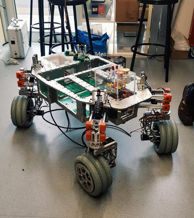 Ein Roboter sitzt auf einem Tisch in einem Raum mit Hockern, Kartons und einem Menschenschuh auf der rechten Seite.