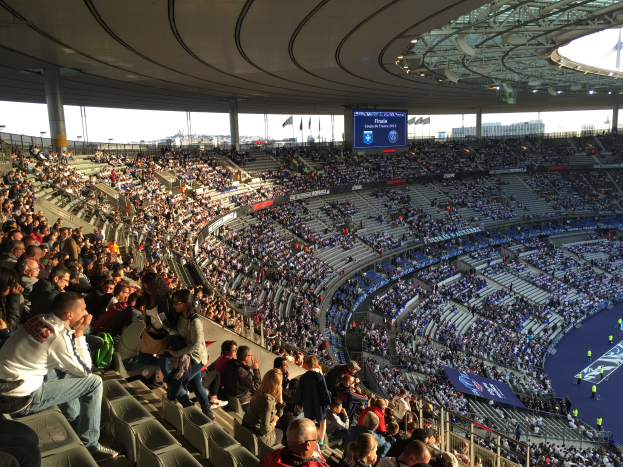 Eine große Menschenmenge sitzt im Allianz Stadion in München, Deutschland, und schaut ein Fußballspiel. Rechts im Bild ist eine Bühne mit ein paar Leuten darauf zu sehen. Im Hintergrund gibt es Fahnen, Stangen und einen Bildschirm. Der Himmel ist oben im Bild zu sehen, und das Stadion scheint das Allianz Stadion in München, Deutschland, zu sein.