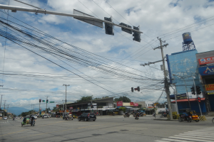 Eine belebte Straße mit zahlreichen Fahrzeugen, Strommästen mit Kabeln, Gebäuden mit Namensschildern, Bäumen und einem bewölktem Himmel.