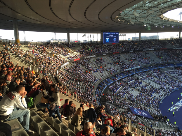 Große Zuschauermenge in einem Stadion bei einem Fußballspiel, mit einer Bühne, Fahnen, Stangen und einem Bildschirm im Hintergrund.