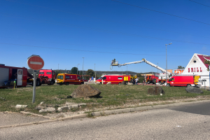 Eine Gruppe von Feuerwehrautos, die auf der Seite einer Straße geparkt sind, umgeben von Gras, Steinen, einem Schild, einem Haus, Pfählen, einem Kran, Drähten, Bäumen und einem bewölkten Himmel, wahrscheinlich nach einem Feuer an einer Tankstelle.