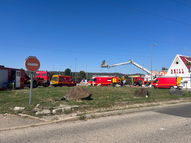 Eine Gruppe von Feuerwehrautos, die auf der Seite einer Straße geparkt sind, umgeben von Gras, Steinen, einem Schild, einem Haus, Pfählen, einem Kran, Drähten, Bäumen und einem bewölkten Himmel, wahrscheinlich nach einem Feuer an einer Tankstelle.
