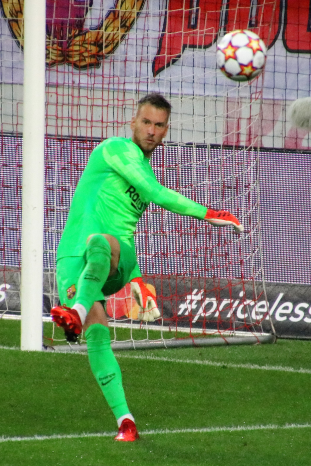 Ein Fußballer in grüner Uniform fängt einen Ball vor einem Tor mit Netz und einem Banner mit Text und Bildern im Hintergrund, trägt Fußballhandschuhe.