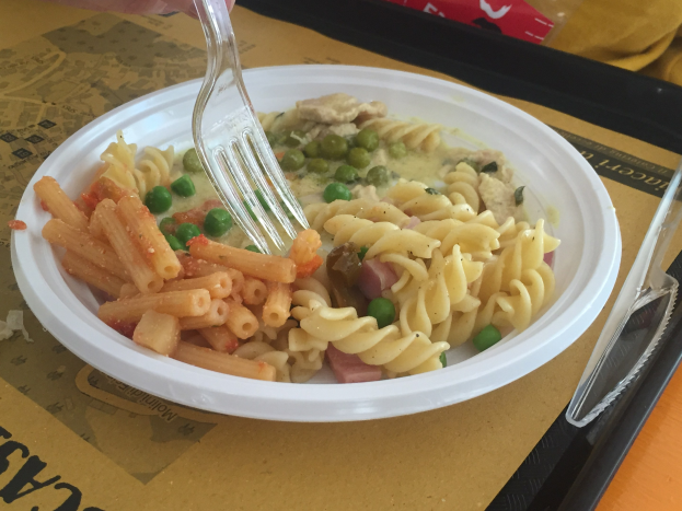 Eine Person, die eine Gabel über eine Schüssel mit Nudeln, Erbsen und Schinken hält, mit einem Tablett, das einen Löffel und eine Abdeckung enthält, auf dem Tisch.