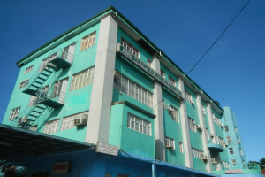 Ein großes grünes Gebäude mit zahlreichen Fenstern, Klimaanlagen und Treppen, umgeben von Bäumen, Kraftfahrzeugen auf der Straße und einem klaren blauen Himmel.