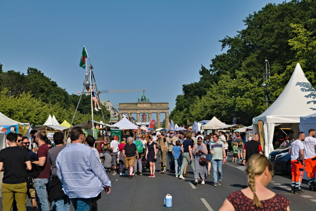 Eine Menge geht eine Straße entlang, die mit Zelten, Fahrzeugen und Bäumen gesäumt ist, auf einen Bogen unter einem klaren blauen Himmel zu, mit Fahnenmästen auf der linken Seite.