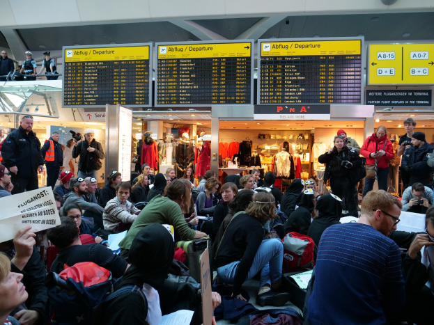 Eine große Gruppe von Menschen versammelt sich in einem Flughafen, einige sitzen mit Taschen und Papieren, andere stehen mit Texttafeln, Schaufensterpuppen in Kleidern und Deckenbeleuchtung im Hintergrund, was auf eine Demonstration hindeutet.