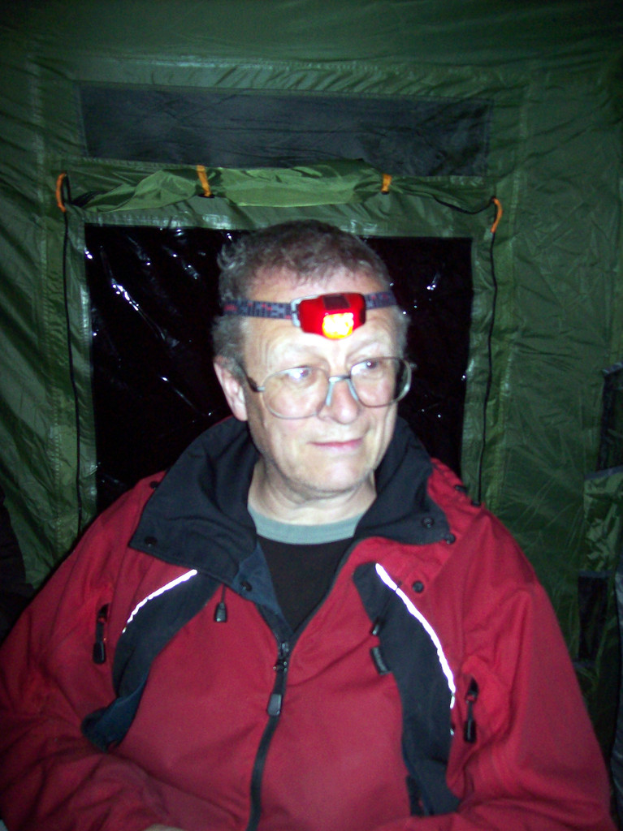 Ein alter Mann in einer roten Jacke ist in einem Zelt, trägt eine Stirnlampen.