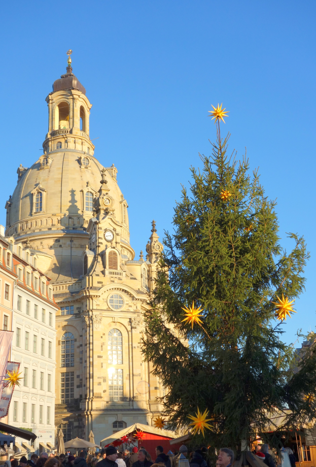 Ein geschäftiger Weihnachtsmarkt in Dresden, Deutschland, mit einem großen Weihnachtsbaum vor einer Kirche, vielen Menschen drumherum und einer Fahne mit Text auf der linken Seite, unter einem sichtbaren Himmel.