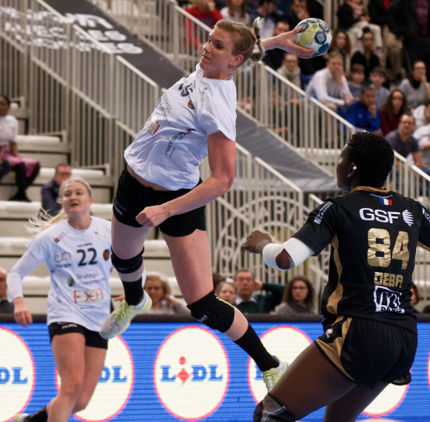 Frauen spielen Handball auf einem Feld mit einem Ball in der Luft, Zuschauern im Hintergrund und einer "Handball-Weltmeisterschaft 2019 - Tag 2"-Tafel.