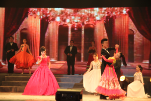 Eine Gruppe von Menschen auf einer Bühne mit Stufen und einem Hintergrundbild, beleuchtet von Scheinwerfern, mit einem Lautsprecher unten, was eine Tanzvorstellung suggeriert.