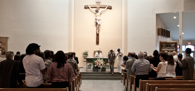 Eine kleine Gruppe Menschen nimmt an einer Hochzeitszeremonie teil, im Hintergrund ist eine Jesus-Statue an einer Wand zu sehen.