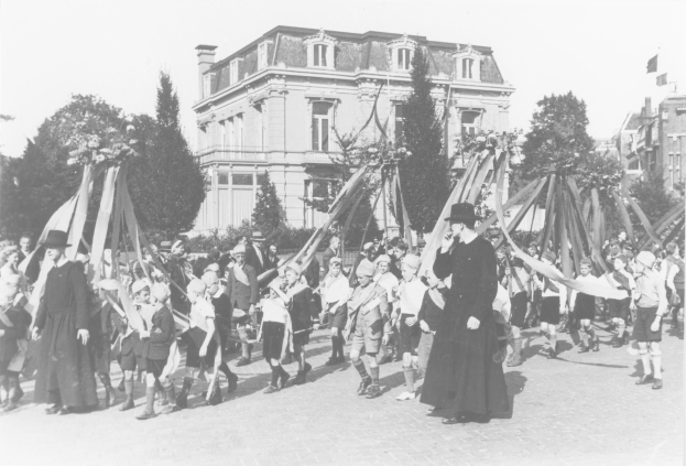 Ein Schwarz-Weiß- Foto von einer Gruppe von Menschen, die mit Fahnen vor einem Gebäude die Straße entlanggehen, mit Bäumen und anderen Gebäuden im Hintergrund und einem Maibaum.