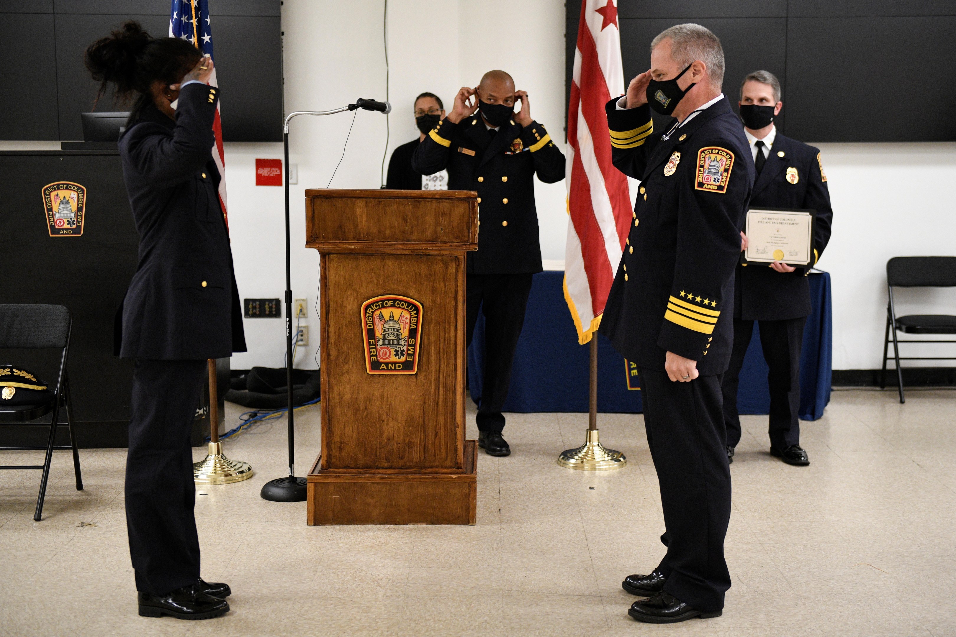 Gruppe uniformierter Personen in Masken auf einem Podium mit einem Mikrofon, umgeben von Flaggen und Möbeln, während einer Vereidigung eines neuen Feuerwehrchefs.