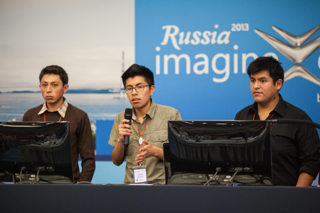 Drei Jungen stehen neben einem Schreibtisch mit zwei Computern, einer spricht in ein Mikrofon und trägt eine ID-Karte mit roter Markierung, vor einer Wand mit einem 'Russia Imagine 2013'-Plakat.