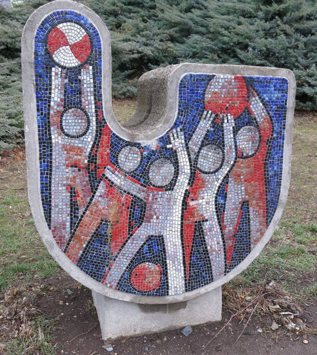 Eine Mosaikbank in einem Park, umgeben von Bäumen unter einem klaren blauen Himmel, mit bunten Fliesen, die ein einzigartiges Design schaffen.