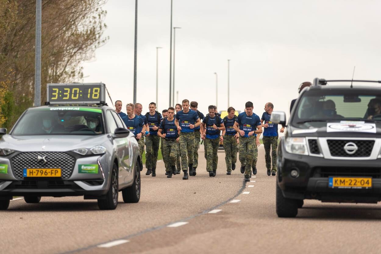 Eine Gruppe von Menschen geht auf einer Straße neben geparkten Autos, mit Bäumen auf der linken Seite und Strommasten im Hintergrund, unter einem sichtbaren Himmel, an einem European Cross Country Challenge teil.