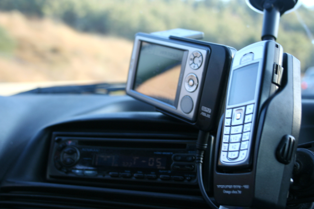 Ein Handy auf einem Mobilhalter mit einer Kamera, einem Kabel und einem MP3-Player auf der linken Seite, ein Glas und unscharfe Objekte im Hintergrund.