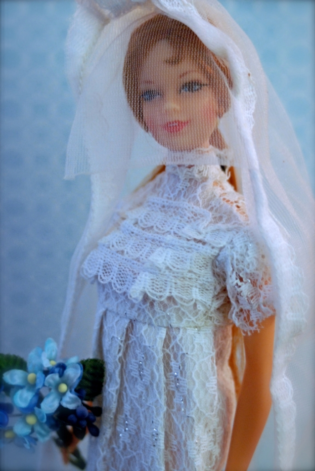 Eine Puppe in Hochzeitskleid mit Schleier und Blumenstrauß, die vor einem unbestimmten Objekt steht.