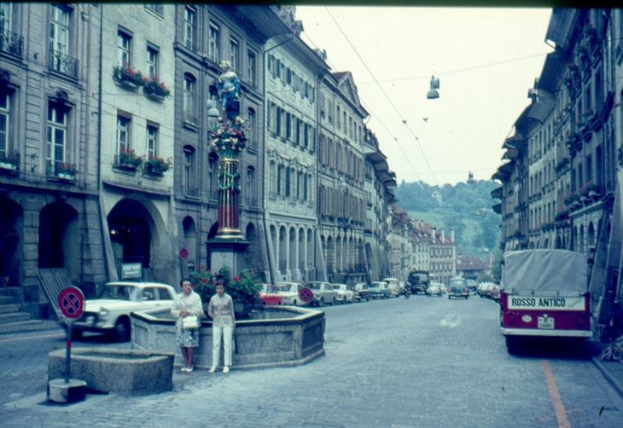 Alte Schwarz-Weiß-Stadtstraße-Szene mit Fußgängern, parkenden Autos, Gebäuden, Fensterblumenkästen, einer Statue auf einem Sockel, einem Wegweiser, einem Fahrzeug auf der Straße, Bäumen und einem klaren blauen Himmel.