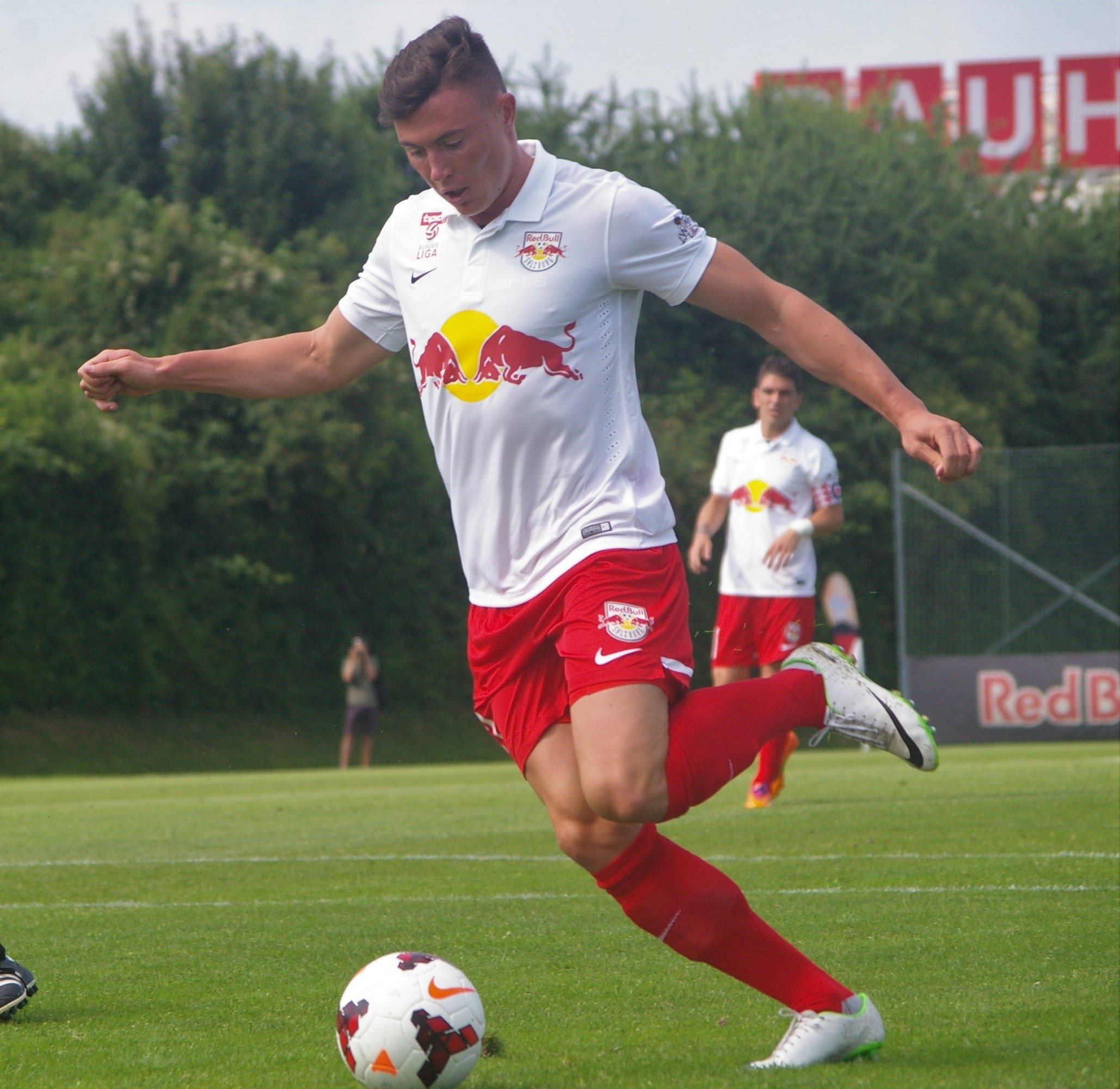 Ein professioneller Fußballspieler, der einen Ball auf einem Rasenfeld mit Bäumen und einem klaren blauen Himmel im Hintergrund kickt, mit einem "RB Leipzig"-Schild im Hintergrund.
