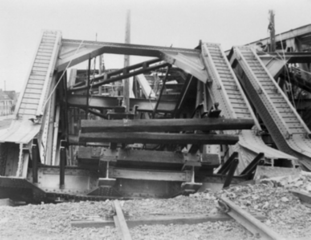 Ein Schwarz-Weiß-Foto einer Bahntrasse, die zu einer baufälligen Brücke mit zerbrochenen Geländern und Schutt führt, umgeben von Gebäuden unter einem klaren Himmel.