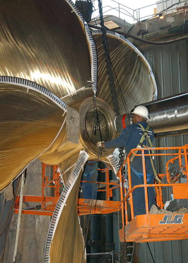 Ein Mann in einem blauen Overall und einem weißen Helm arbeitet an einer großen Turbine in einer Fabrik mit einem orangen Hubsteiger, mit großen Metallrohren und Eisenstangen im Hintergrund.