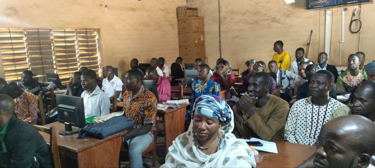Eine Gruppe von Menschen, die an Tischen in einem Klassenzimmer sitzen, mit Monitoren, Taschen, Büchern, Papieren und anderen Gegenständen auf den Tischen, einem Fenster auf der linken Seite, Kartons und einem Fernseher an der Wand, an einer Techniktrainingssitzung teilnehmen.