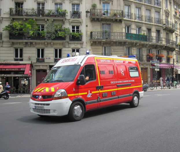 Ambulanz auf der Seite einer Straße vor einem Gebäude mit Fenstern, Geländern, Pflanzen und anderen Gegenständen geparkt, mit einigen Menschen und Fahrrädern im Hintergrund.