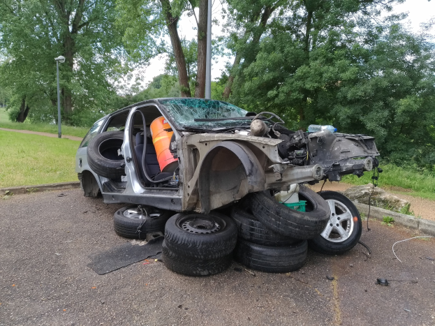 Ein umgestürzter Wagen am Straßenrand mit verstreuter Trümmer, umgeben von Gras, Pflanzen, Bäumen, einem Laternenpfahl und einem bewölkten Himmel.