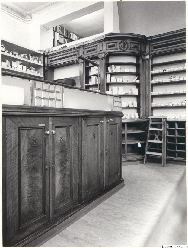 Schwarz-weiß-Foto eines vintage Apothekeninneren mit einer Theke, Schränken, einer Leiter, Regalen und Text oben.