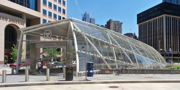 Belebte Stadtstraße mit hohen Wolkenkratzern, Bushaltestelle mit Glasdach, Fußgänger, Fahrzeuge, Metallzaun, Mülltonne, Strommasten, ein Schild, eine Ampel, Bäume, ein Baukran und ein bewölkter Himmel.
