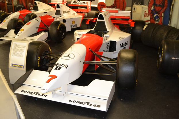 Rennwagen in einem Museum ausgestellt, mit Besuchern, Informationsschild, Absperrseil und Wand im Hintergrund.