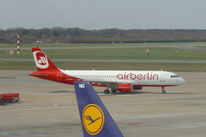 Ein Air-Berlin-Airbus A320-200 auf der Rollbahn am Frankfurter Flughafen, umgeben von Fahrzeugen auf der linken Seite und Gras am Boden. Im Hintergrund sind Poles, Bäume und ein klarer blauer Himmel zu sehen.