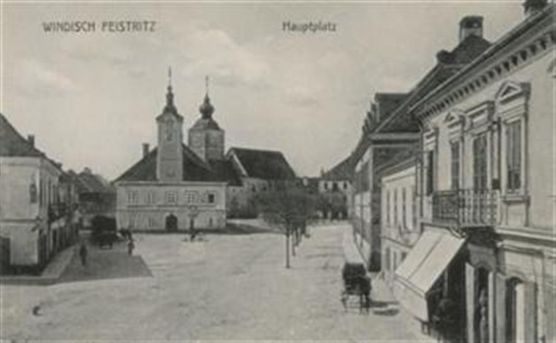 Ein altes Schwarz-Weiß-Foto einer Stadtstraße in Windisch Feststritz, Deutschland, mit Gebäuden, Bäumen, Menschen, Karren und einem Himmel im Hintergrund, mit Text oben und unten auf dem Bild.