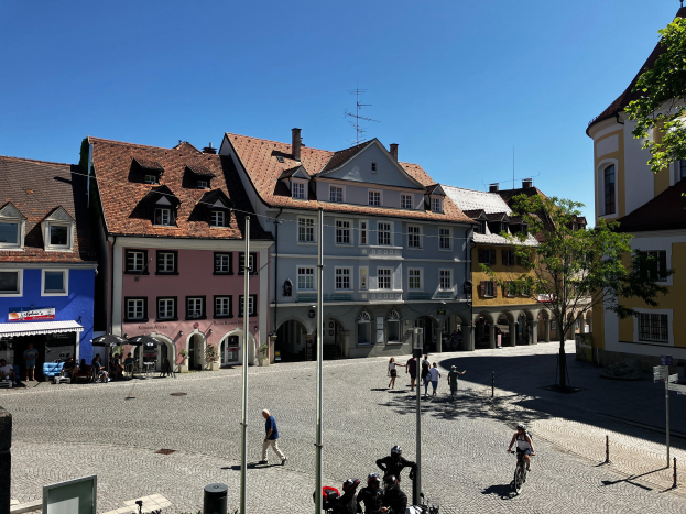 Eine Gruppe von Menschen, die auf Fahrrädern eine Kopfsteinpflasterstraße in der Altstadt von Heidelberg entlangfahren, mit Gebäuden, Laternenpfählen, Bäumen und Bänken im Hintergrund, unter einem sichtbaren Himmel.