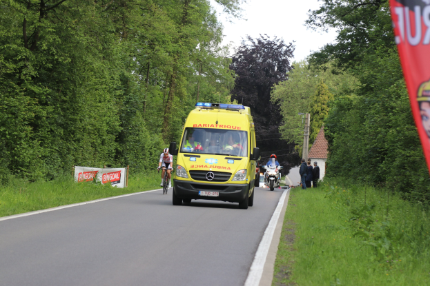 Ambulanz fährt auf Straße mit Fahrradfahrern daneben, Gras und Bäume auf beiden Seiten, Häuser und Masten im Hintergrund unter einem klaren blauen Himmel.