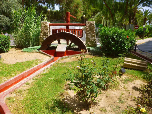 Minigolf-Platz in einem Park mit Grünfläche, einer Straße mit Fußgängern, Schildern, Bäumen und einem klaren Himmel.