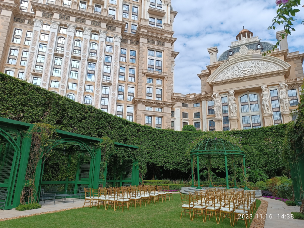 Eine Trauungszeremonie findet vor einem großen Gebäude mit Glasfenstern statt, umgeben von üppiger Vegetation, mit Stühlen auf dem Boden und einem Pavillon im Vordergrund, unter einem klaren blauen Himmel.