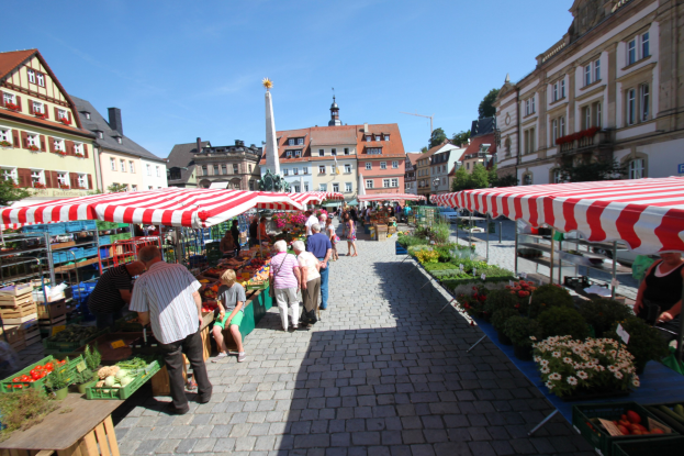 Ein belebter Markt im historischen Zentrum von Heidelberg mit Menschen, die spazieren gehen, auf Bänken sitzen und in der Nähe von Zelten stehen, mit Gemüsekörben auf Tischen, Gebäuden mit Fenstern, Bäumen und einem klaren blauen Himmel im Hintergrund.