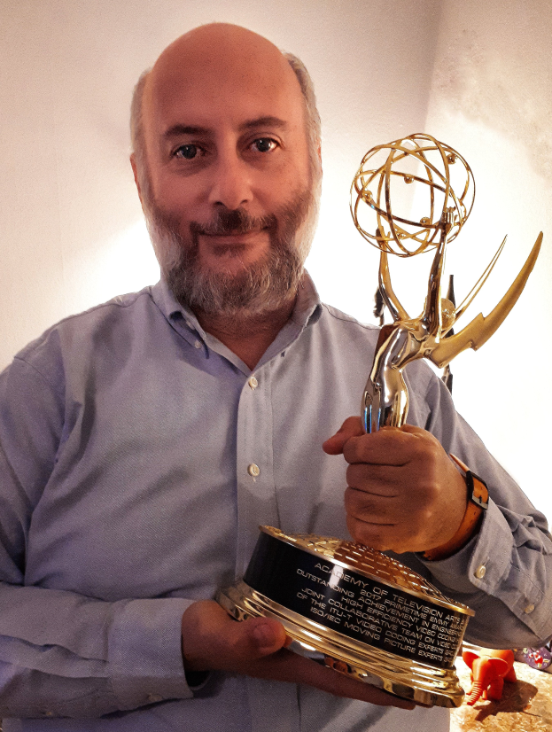 Ein Mann mit einem Emmy Award in der Hand und einem Lächeln im Gesicht, mit Gegenständen auf einem Tisch und einer Wand im Hintergrund.