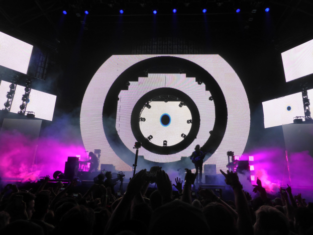 Menschenmenge auf einem Konzert mit erhobenen Händen, einige halten Telefone oder spielen Instrumente, großer Bildschirm und Lichter im Hintergrund.