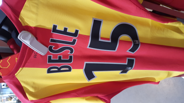 Ein rotes und gelbes Fußballtrikot mit Text und Zahlen darauf, das an einer Stange in einem Geschäft hängt, zusammen mit anderen Gegenständen und dem Boden im Hintergrund.