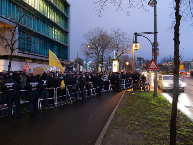 Große Gruppe von Menschen vor einem Gebäude in Berlin mit Protestschildern, Barrikaden, Fahrrädern, Schildern, Bäumen und Gras im Vordergrund.